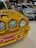 A close-up view of a vintage yellow race car with prominent rally headlights and a red license plate. The car's hood features branding and artwork, and it is parked in an indoor showroom alongside other racing cars. Another car seen in the background has a silver and blue color scheme with various logos.