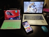 A desk setup featuring three electronic devices. An HP laptop with a screen showing a woman hugging a lion is placed next to a tablet with a green backlit keyboard and a car wallpaper on its display. A smartphone with colorful app icons and a vehicle image on the lock screen rests in front of them. A blue wireless mouse and a wristwatch are also visible on the desk.