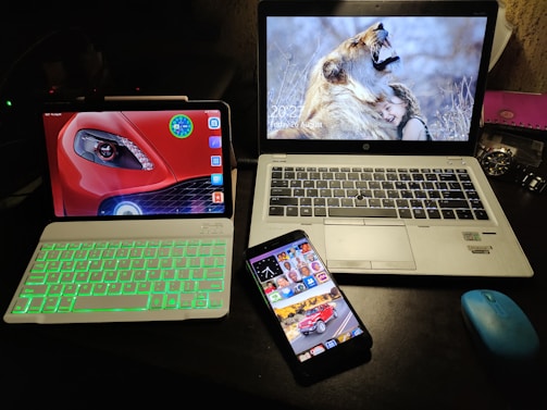 A desk setup featuring three electronic devices. An HP laptop with a screen showing a woman hugging a lion is placed next to a tablet with a green backlit keyboard and a car wallpaper on its display. A smartphone with colorful app icons and a vehicle image on the lock screen rests in front of them. A blue wireless mouse and a wristwatch are also visible on the desk.
