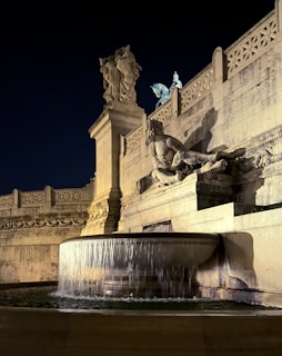 An ornate stone sculpture of a reclining figure is prominently displayed alongside a cascading water fountain. The scene is illuminated against a deep blue night sky, with intricate architectural details and additional statues visible on the structure behind the main sculpture.