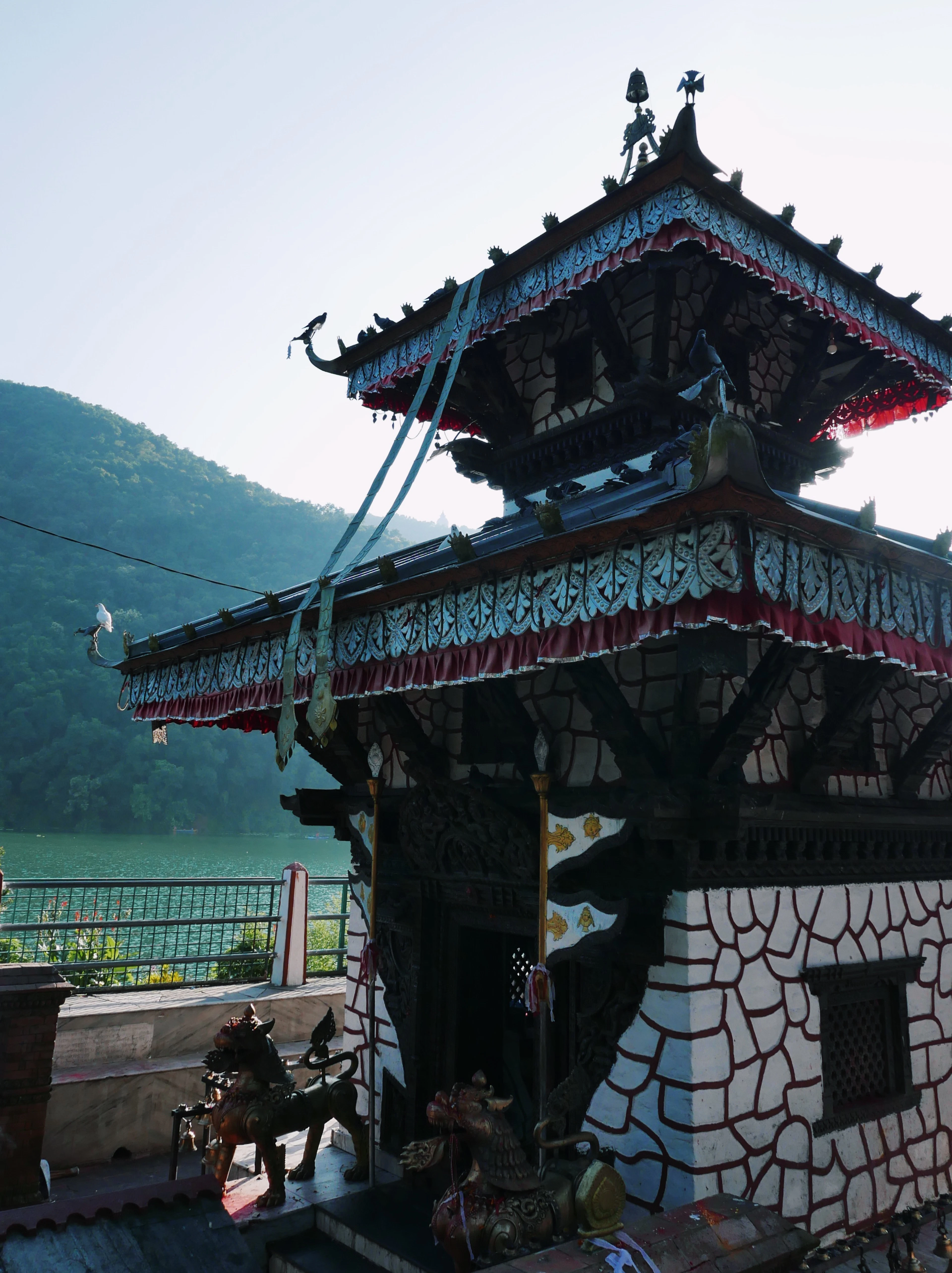 The two-tiered pagoda of Tal Barahi temple on its island in Phewa Lake, its carved eaves and red-and-white shrine walls close up, with brass guardian lions at the steps and the wooded far shore beyond the water