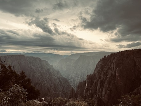 A dramatic landscape featuring deep canyons and rugged cliffs under a sky filled with dark, swirling clouds. The lighting creates a moody atmosphere, with the distant mountains barely visible through the misty haze.