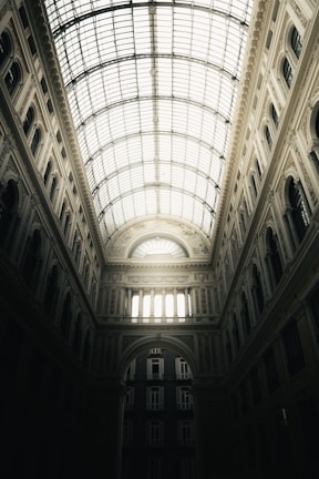 A vast and ornate interior space is illuminated by a large, arched glass ceiling, casting light onto the intricate architectural details. The walls are adorned with decorative molding, and arched windows are symmetrically aligned along the structure. The design is grand and imposing, with light filtering through to reveal the elegance of the crafted elements.