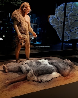 A life-sized display of what appears to be two prehistoric human figures. One figure, standing, is covered in fur clothing, while another lies on the ground, also draped in furs, appearing to be at rest or sleeping. The setting suggests a primitive environment, with a dimly lit background and structures that resemble a museum exhibit.