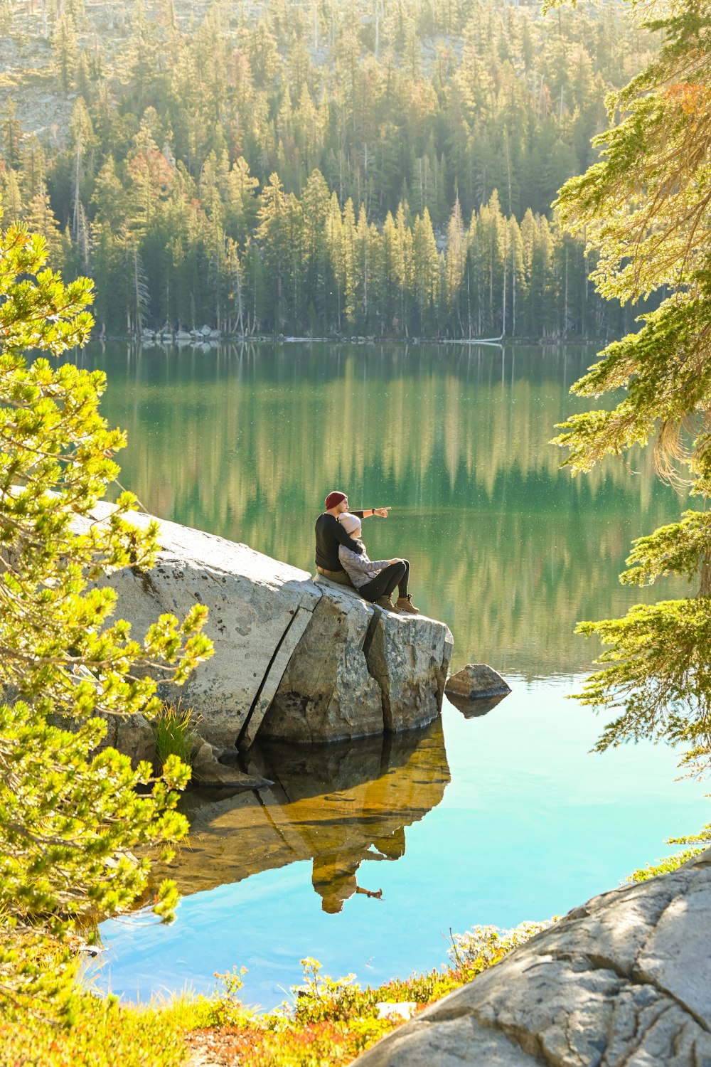 a person sitting on a rock by a lake with trees and hills in the background