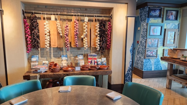 A cozy interior space with a rustic theme, featuring a round table with green cushioned chairs and napkins. The wall showcases decorative strings of garlic and chili peppers hanging on cutting boards. Below them, on a wooden table, are terracotta dishes and various packaged items. The wall is adorned with patterned wallpaper and framed photos.