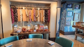A cozy interior space with a rustic theme, featuring a round table with green cushioned chairs and napkins. The wall showcases decorative strings of garlic and chili peppers hanging on cutting boards. Below them, on a wooden table, are terracotta dishes and various packaged items. The wall is adorned with patterned wallpaper and framed photos.