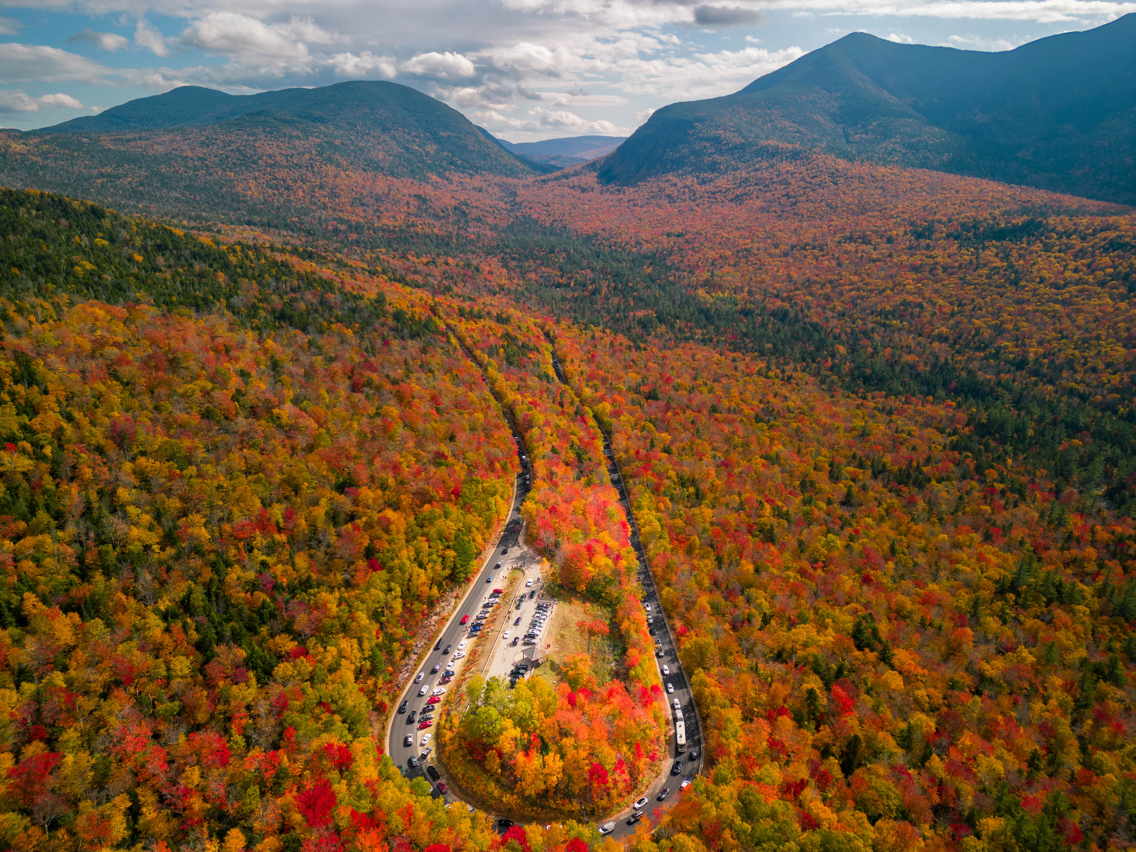 The Kancamagus Highway in New Hampshire from above: a bend through forest turned red and gold, with the White Mountains beyond