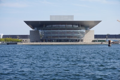 A modern architectural building with a sleek, expansive roof overhangs the structure, located by a large body of water. The facade consists of a combination of glass and stone elements, with multiple stories visible. In the background, a long, dark-roofed building is partially covered by greenery. The sky is clear and blue.