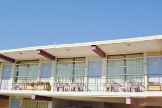 A two-story motel facade is depicted with a flat roof and a series of windows and balconies. Each balcony is furnished with a small table and a pair of chairs. Window boxes filled with vibrant flowers decorate the railing. The building features a color scheme of white and purple, and the sky is clear and blue in the background.