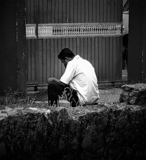 A person in a white shirt is sitting alone on a stone ledge with their head bowed and arms resting on their knees. The background features a large, ornate metal gate. The scene is depicted in black and white, giving it a nostalgic and somber feel.