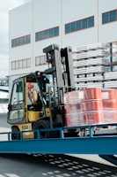 A forklift operator maneuvering a yellow forklift carrying several stacked, red barrels wrapped in plastic. The scene is set outside a warehouse-like building with multiple windows. Metal racks with stored items are visible in the background.