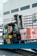 A forklift operator maneuvering a yellow forklift carrying several stacked, red barrels wrapped in plastic. The scene is set outside a warehouse-like building with multiple windows. Metal racks with stored items are visible in the background.