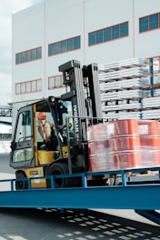 A forklift operator maneuvering a yellow forklift carrying several stacked, red barrels wrapped in plastic. The scene is set outside a warehouse-like building with multiple windows. Metal racks with stored items are visible in the background.