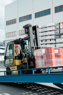 A forklift operator maneuvering a yellow forklift carrying several stacked, red barrels wrapped in plastic. The scene is set outside a warehouse-like building with multiple windows. Metal racks with stored items are visible in the background.