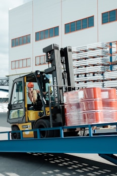 A forklift operator maneuvering a yellow forklift carrying several stacked, red barrels wrapped in plastic. The scene is set outside a warehouse-like building with multiple windows. Metal racks with stored items are visible in the background.
