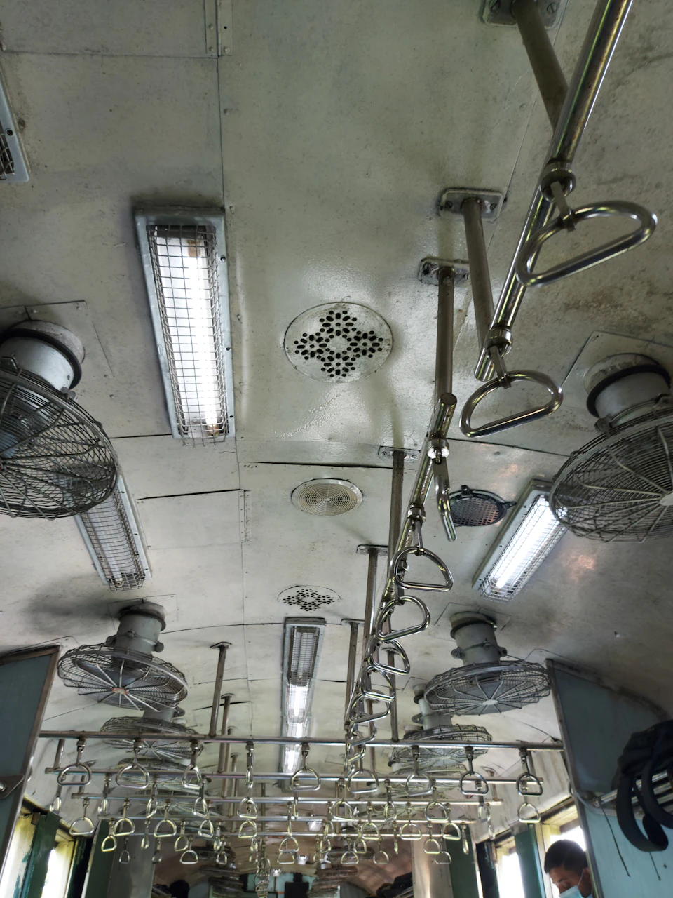 The image shows a train car interior with white walls and ceiling, illuminated by multiple lights that cast a soft glow throughout the space. The ceiling features a grid-like pattern composed of small square panels, adding an architectural element to the room. Several hanging fans are present in various positions, providing ventilation for passengers during their journey.