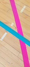 Brightly colored lines of pink and blue are taped across a wooden floor. Several off-white tape pieces are also visible, likely serving as markers.