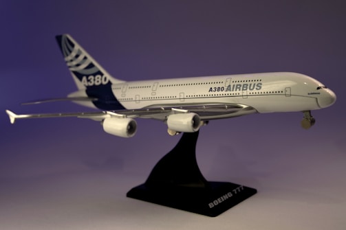 A model airplane of the Airbus A380 is displayed on a black stand. The aircraft is white with blue accents, featuring detailed engines and landing gear. The lighting casts a gradient of purple hues in the background.