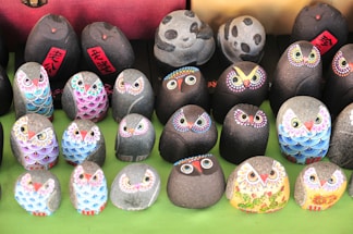 A collection of small, intricately painted stones arranged in rows. Each stone is decorated with vibrant colors and features that resemble owls, with detailed patterns on their faces and feathers. The stones have a smooth texture and are placed on a green surface. Some stones feature additional Chinese characters in red.