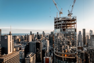 A high-rise building under construction dominates the foreground of an urban skyline, with cranes perched atop while a multitude of completed skyscrapers stretches into the horizon beneath a blue sky.