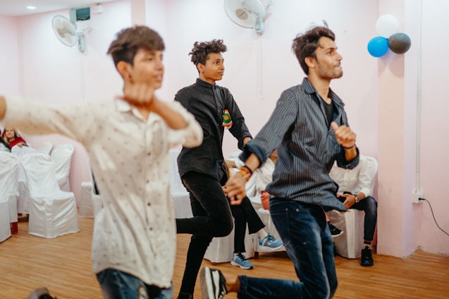 Three young men are energetically dancing in a room with light pink walls. White chairs covered with cloth are arranged along the sides. A cluster of blue, black, and white balloons is visible on the wall, suggesting a celebratory event. Ceiling fans are installed on the walls, and the wooden floor adds warmth to the setting.