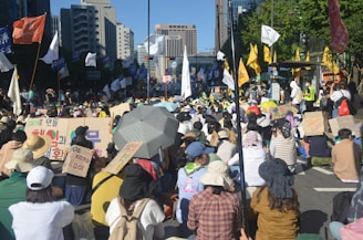 A large crowd of people is gathered outdoors on a city street. Many individuals are seated, holding signs with text in various languages. Flags of different colors are visible, and a few people are standing. The scene has an urban backdrop with tall buildings and trees.
