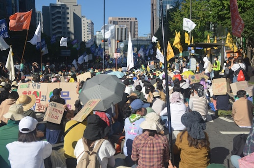 A large crowd of people is gathered outdoors on a city street. Many individuals are seated, holding signs with text in various languages. Flags of different colors are visible, and a few people are standing. The scene has an urban backdrop with tall buildings and trees.