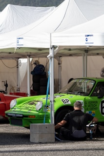 A person is seated and appears to be working on or near a bright green race car under a white tent. The car has various decals and sponsor stickers, including the letters 'BP'. There are additional items such as a red car next to the green one, stacked tires, and other equipment visible in the background.