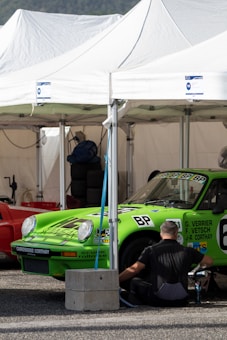 A person is seated and appears to be working on or near a bright green race car under a white tent. The car has various decals and sponsor stickers, including the letters 'BP'. There are additional items such as a red car next to the green one, stacked tires, and other equipment visible in the background.