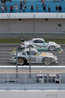 Two race cars positioned on a track near a barrier are preparing for a race. Spectators are gathered in the stands watching and taking photos. The cars have various sponsorship logos, including Fujifilm, displayed prominently. The environment appears competitive and energetic.