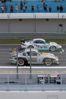 Two race cars positioned on a track near a barrier are preparing for a race. Spectators are gathered in the stands watching and taking photos. The cars have various sponsorship logos, including Fujifilm, displayed prominently. The environment appears competitive and energetic.