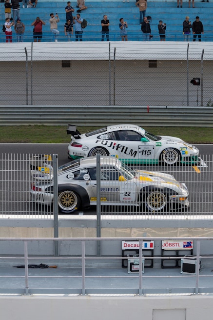 Two race cars positioned on a track near a barrier are preparing for a race. Spectators are gathered in the stands watching and taking photos. The cars have various sponsorship logos, including Fujifilm, displayed prominently. The environment appears competitive and energetic.