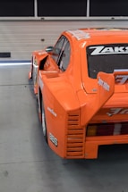 An orange sports car with distinctive body styling is parked indoors. The back part of the car is visible, showing branding and logos such as Goodyear and BBS on the side. The car features a prominent rear spoiler and the name 'ZAKS' on the rear window.