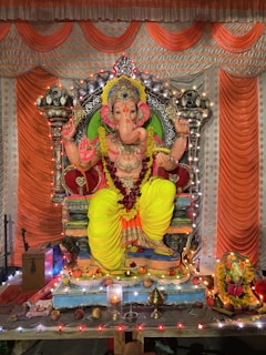 A detailed idol of Lord Ganesha is adorned with vibrant decorations and intricate jewelry. The statue is seated on an ornate throne with vivid yellow and orange drapery. Surrounding the idol are various offerings such as fruits and flowers. The setting is decorated with strings of lights, enhancing the festive atmosphere.
