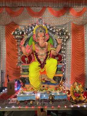 A detailed idol of Lord Ganesha is adorned with vibrant decorations and intricate jewelry. The statue is seated on an ornate throne with vivid yellow and orange drapery. Surrounding the idol are various offerings such as fruits and flowers. The setting is decorated with strings of lights, enhancing the festive atmosphere.