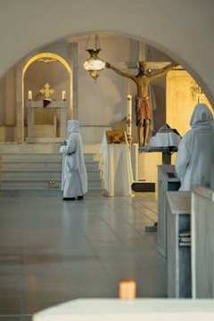 Two figures in white robes stand in a dimly lit chapel. One is near an altar holding a book, and the other is closer to a crucifixion statue of Jesus. The setting is serene, with candlelight adding to the tranquil atmosphere.
