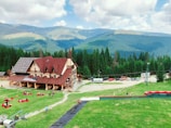 A scenic mountain view with a large rustic wooden building featuring solar panels on the roof, surrounded by lush greenery and a forest of coniferous trees. People are relaxing on bean bags spread across the grassy area in front of the building, and several cars are parked nearby. Rolling mountains are visible in the background under a partly cloudy sky.