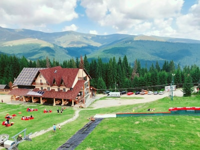 A scenic mountain view with a large rustic wooden building featuring solar panels on the roof, surrounded by lush greenery and a forest of coniferous trees. People are relaxing on bean bags spread across the grassy area in front of the building, and several cars are parked nearby. Rolling mountains are visible in the background under a partly cloudy sky.