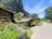 A picturesque village scene with wooden houses surrounded by vibrant gardens full of colorful flowers. A large rocky cliff looms in the background, providing a dramatic backdrop. Several people are gathered under a small wooden shelter, and a pathway leads through the scene under a clear blue sky.
