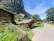 A picturesque village scene with wooden houses surrounded by vibrant gardens full of colorful flowers. A large rocky cliff looms in the background, providing a dramatic backdrop. Several people are gathered under a small wooden shelter, and a pathway leads through the scene under a clear blue sky.