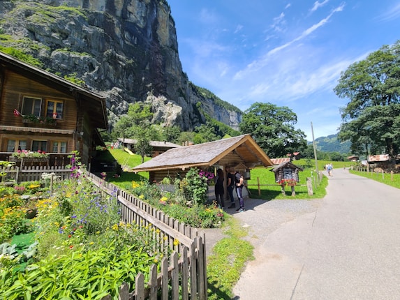 A picturesque village scene with wooden houses surrounded by vibrant gardens full of colorful flowers. A large rocky cliff looms in the background, providing a dramatic backdrop. Several people are gathered under a small wooden shelter, and a pathway leads through the scene under a clear blue sky.