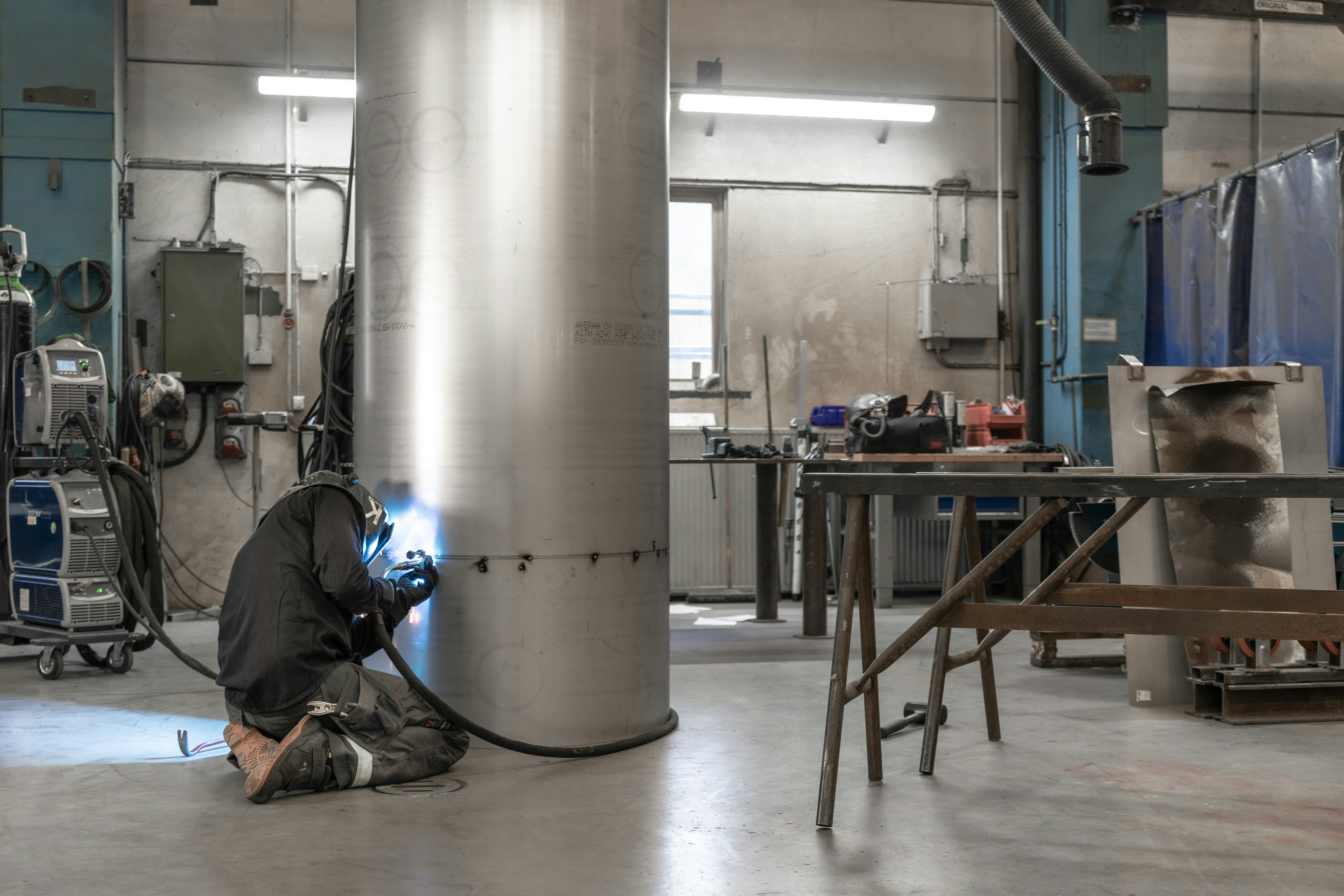 a person working in a factory, A welder welding on a big metallic tube.