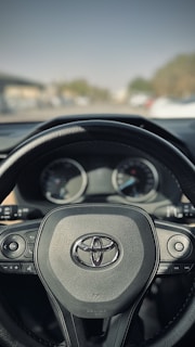A close-up view of a car steering wheel featuring the Toyota logo in the center. The steering wheel has various buttons for controlling car functions, and the background shows a blurred view of the road and some greenery.