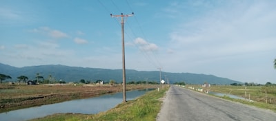 A rural road stretches into the distance, flanked by grassy fields and irrigation ditches. Power lines run alongside the road with a backdrop of distant mountains under a bright blue sky. Scattered trees and small buildings are visible on the horizon.