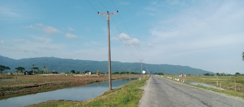 A rural road stretches into the distance, flanked by grassy fields and irrigation ditches. Power lines run alongside the road with a backdrop of distant mountains under a bright blue sky. Scattered trees and small buildings are visible on the horizon.