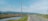 A rural road stretches into the distance, flanked by grassy fields and irrigation ditches. Power lines run alongside the road with a backdrop of distant mountains under a bright blue sky. Scattered trees and small buildings are visible on the horizon.