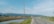 A rural road stretches into the distance, flanked by grassy fields and irrigation ditches. Power lines run alongside the road with a backdrop of distant mountains under a bright blue sky. Scattered trees and small buildings are visible on the horizon.