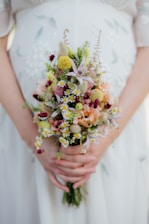 A person is holding a floral bouquet with a variety of small, colorful flowers including daisies, yellow billy balls, and pink blossoms. The hands gently clasp the stems, and the background shows a white dress with subtle embroidery.