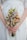 A person is holding a floral bouquet with a variety of small, colorful flowers including daisies, yellow billy balls, and pink blossoms. The hands gently clasp the stems, and the background shows a white dress with subtle embroidery.